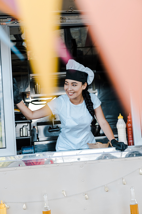 selective focus of cheerful asian woman in hat and chef uniform smiling in food truck