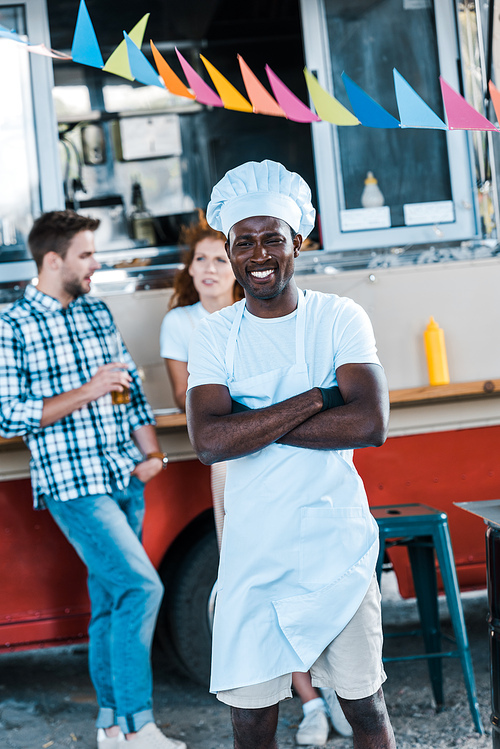selective focus of happy african american chef standing with crossed arms near food truck and customers