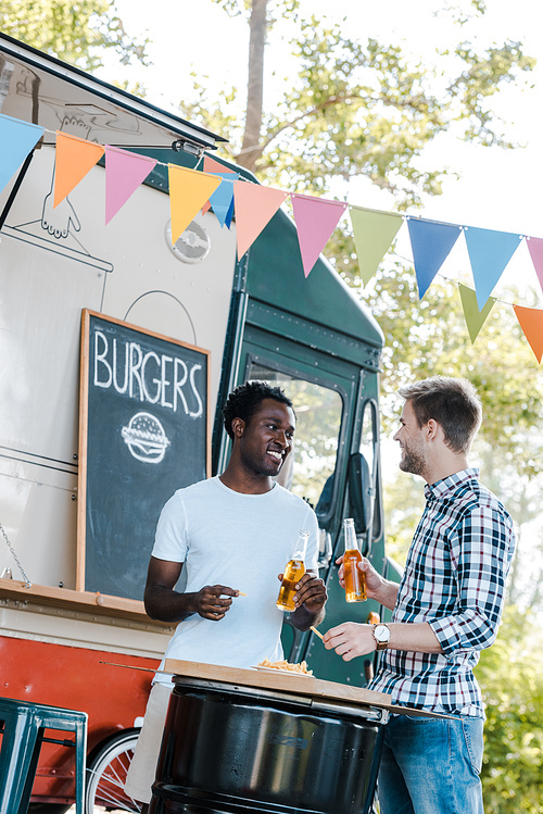 cheerful multicultural friends holding bottles of beer near french fries and food truck