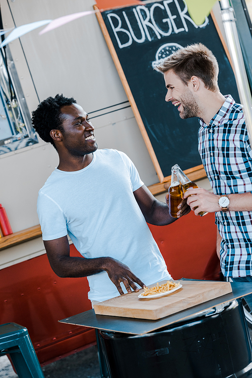 positive multicultural men toasting bottles of beer near french fries and food truck