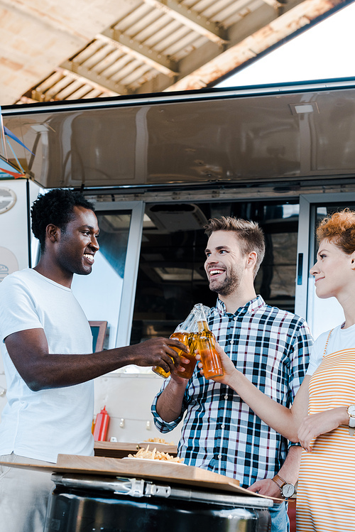 selective focus of multicultural men clinking bottles of beer with happy redhead woman near food truck