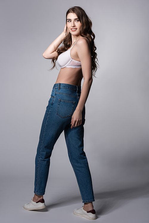 cheerful girl in bra and blue jeans standing on grey