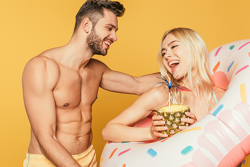 smiling muscular man touching excited girl in swim ring holding pineapple with cocktail isolated on yellow