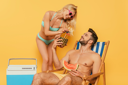 cheerful blonde girl giving pineapple with cocktail to handsome man sitting in deck chair with slice of watermelon on yellow background