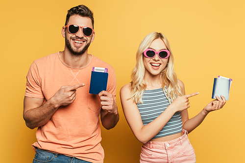 happy young couple in sunglasses pointing with fingers at passports and air tickets on yellow background