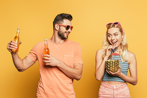 smiling man holding bottles of beer near cheerful girl drinking cocktail from pineapple on yellow background
