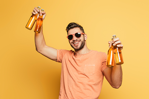 cheerful young man in sunglasses holding bottles of beer while smiling at camera on yellow background