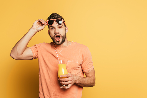 shocked young man with open mouth touching sunglasses while holding orange juice on yellow background