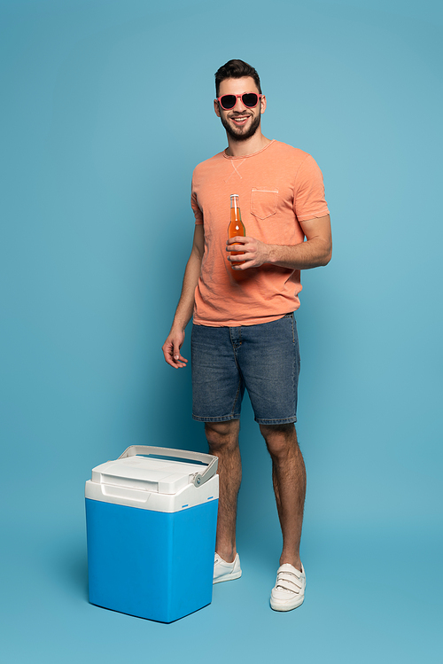happy man in sunglasses holding bottle of beer while standing near portable fridge on blue background