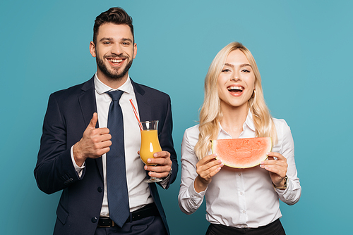 happy businessman with orange juice showing thump up near smiling businesswoman with juicy watermelon on blue background