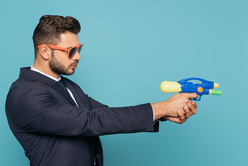 side view of businessman in sunglasses holding toy water gun isolated on blue