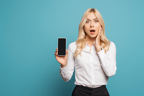 shocked businesswoman touching face while holding smartphone with blank screen on blue background