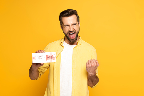 Cheerful man showing yeah gesture and holding gift card on yellow background