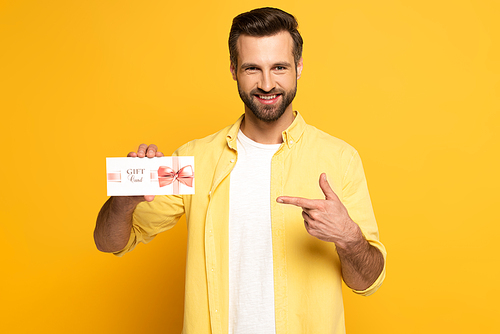 Smiling man pointing with finger at gift card on yellow background