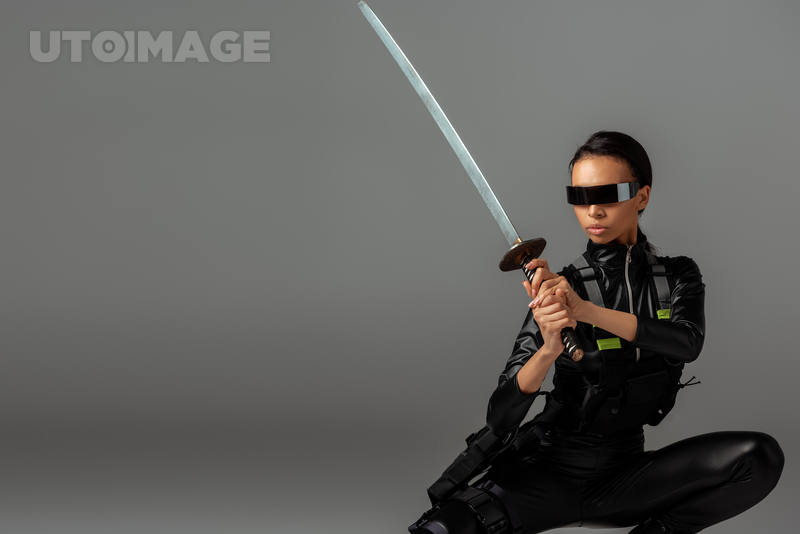 유토이미지 | attractive futuristic african american woman in glasses sitting with sword on grey
