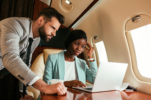 two multicultural businesspeople looking at laptop while traveling in private jet