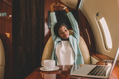 smiling african american businesswoman stretching with raised hands near laptop and cup of coffee in private jet