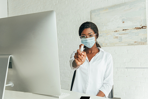 young african american freelancer in medical mask showing antiseptic spray while sitting near computer monitor at home