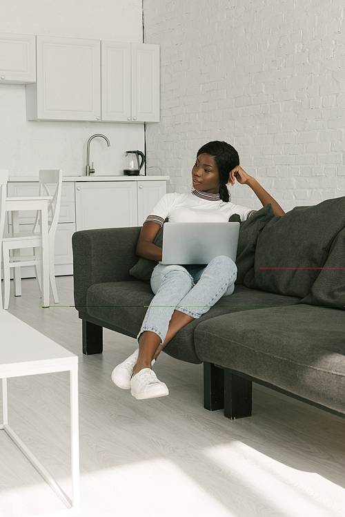 pensive african american freelancer sitting on sofa with laptop and looking away