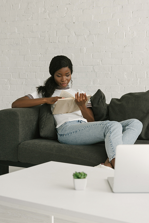 young african american freelancer resting on sofa while reading book near table with laptop