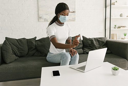 young african american freelancer in medical mask spraying antiseptic on laptop