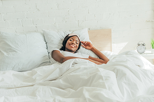 happy african american girl with sleep mask on forehead lying in bed and smiling at camera