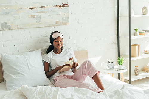 smiling african american girl with sleep mask on forehead sitting in bed, holding book and 