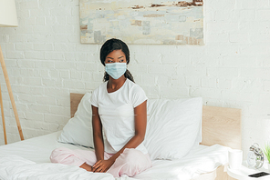 dreamy african american woman in medical mask sitting in bed with crossed legs and looking away