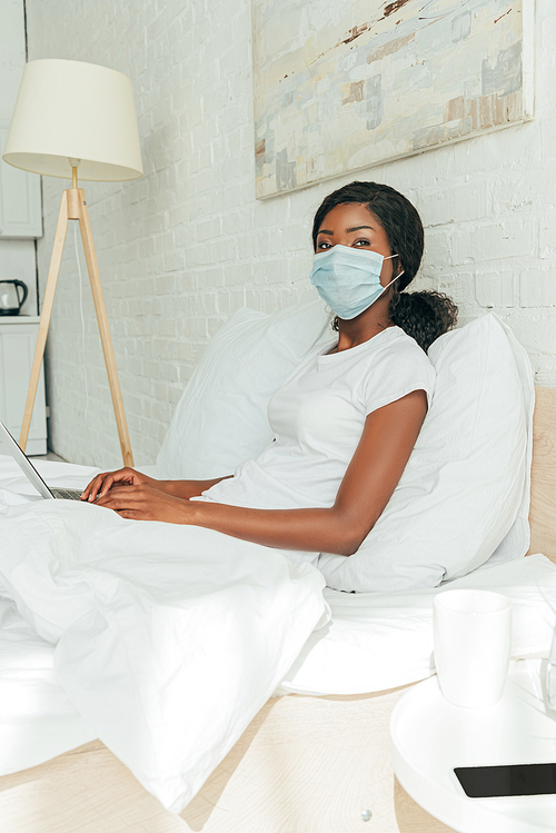 african american freelancer in medical mask sitting in bed with laptop and