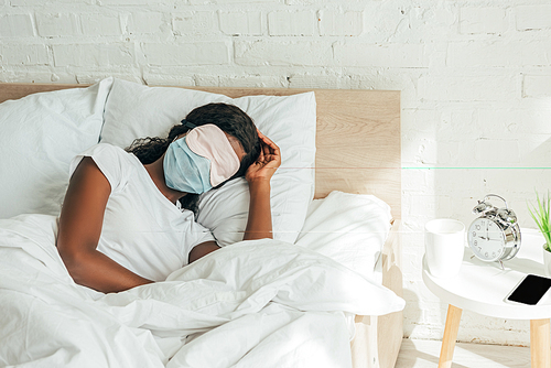 african american woman in sleep mask and medical mask sleeping on white bedding