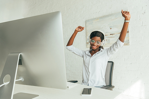 happy african american freelancer gesturing with raised hands while listening music in wireless headphones near computer monitor
