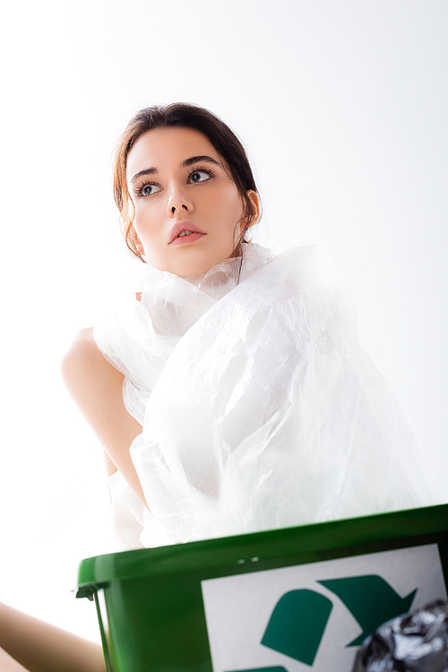 brunette woman wrapped in plastic bag sitting near trash bin with recycle sign isolated on white, ecology concept