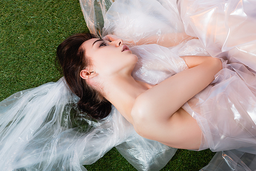 top view of young woman wrapped in polyethylene lying on fresh grass, ecology concept