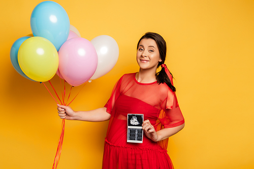 pregnant woman in red outfit holding colorful festive balloons while showing ultrasound scan on yellow