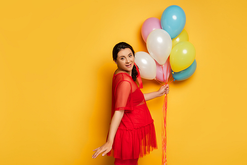 pregnant woman in red tunic holding colorful festive balloons while  on yellow