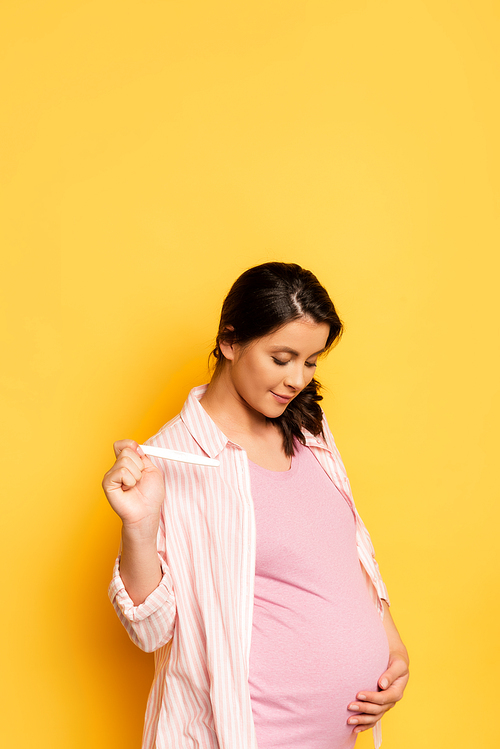 pregnant young woman holding pregnancy test while touching tummy on yellow