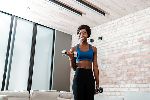 African american sportswoman smiling, training with dumbbells and  in living room