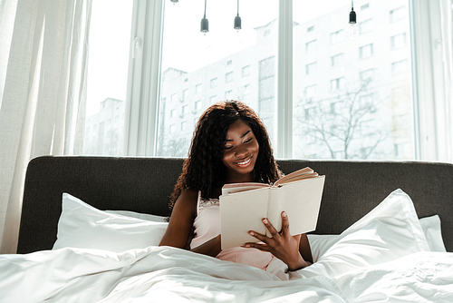 African american woman reading book and smiling on bed in bedroom