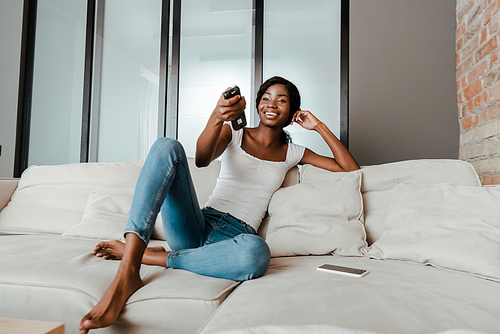 African american woman smiling with remote controller near smartphone on sofa in living room