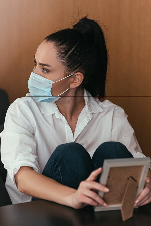 depressed young woman in medical mask looking away while holding photo frame