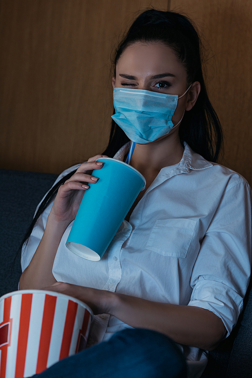 young woman in medical mask winking at camera while holding soda and popcorn