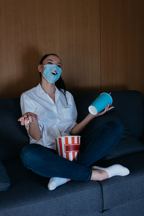 smiling young woman in medical mask with hole watching tv with soda and popcorn at home