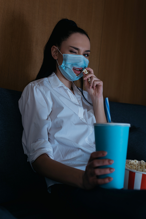 selective focus of young woman in medical mask with hole winking at camera while holding soda and eating popcorn