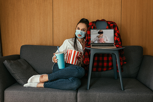 young woman in medical mask with hole holding popcorn and soda near laptop with boyfriend in vr headset on screen