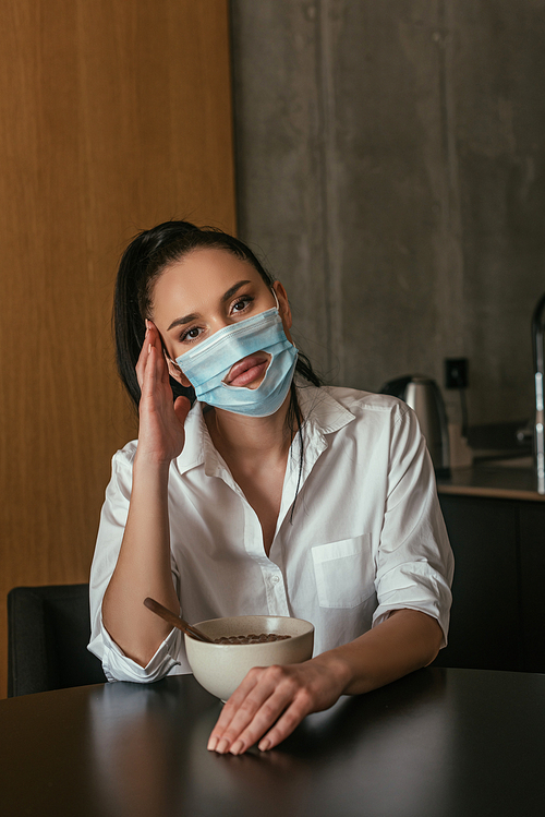 depressed woman in medical mask with hole  while sitting near bowl with breakfast