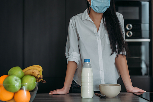 cropped view of woman in protective mask near bottle of milk, bowl and fresh fruits in kitchen