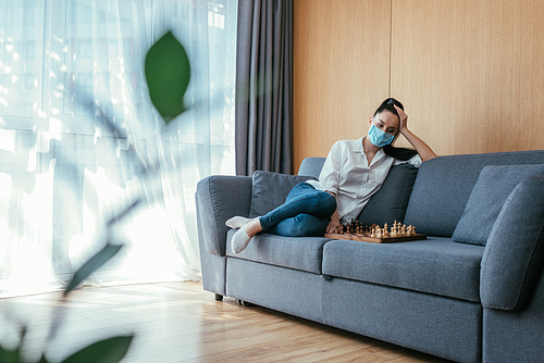 selective focus of sad woman in medical mask sitting near chessboard on sofa