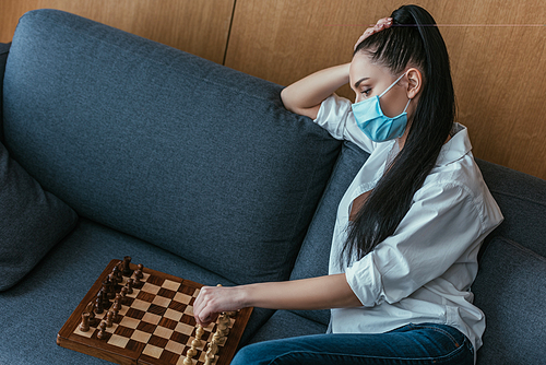 sad woman in medical mask sitting on sofa near chessboard