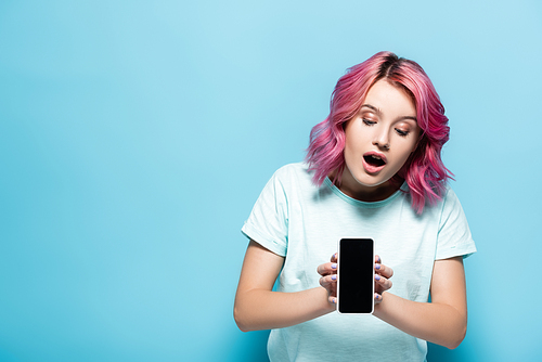 shocked young woman with pink hair holding smartphone with blank screen on blue background