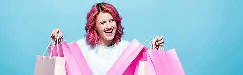 excited young woman with pink hair holding shopping bags isolated on blue , panoramic shot